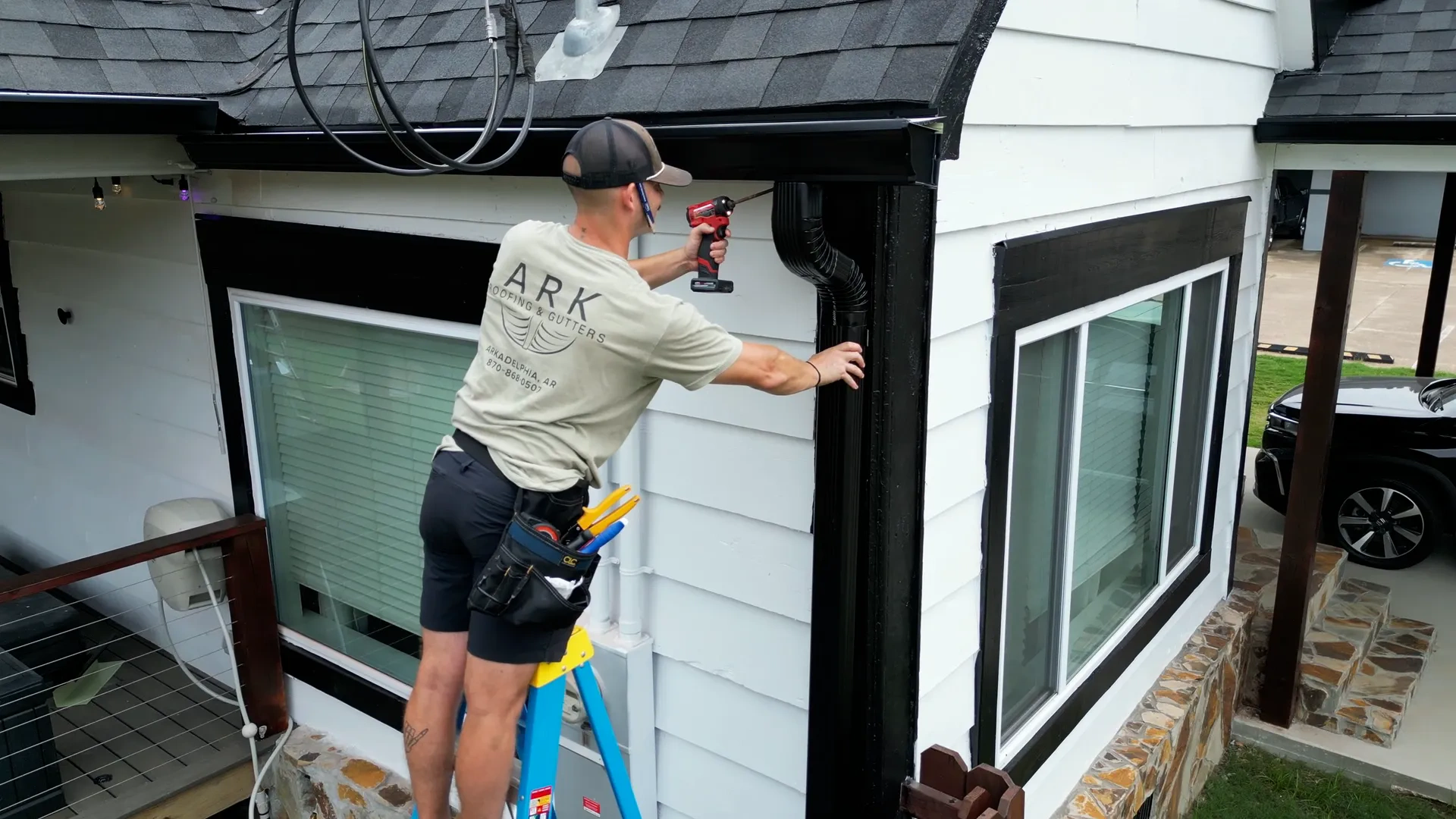 Ark Roofing and Gutters crew member in a branded shirt securing a downspout to the wall of an Arkadelphia home