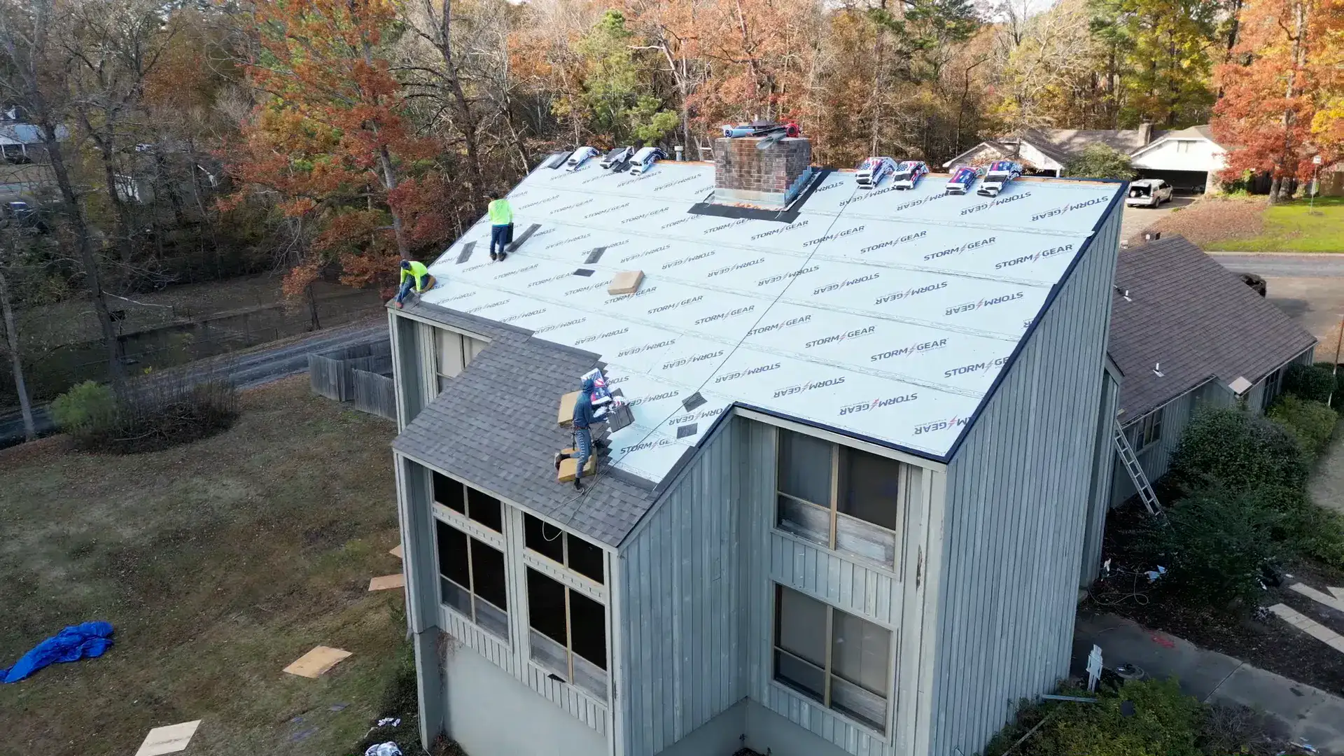 Crew rolling underlayment across a large wooded-lot roof buried in autumn leaf litter