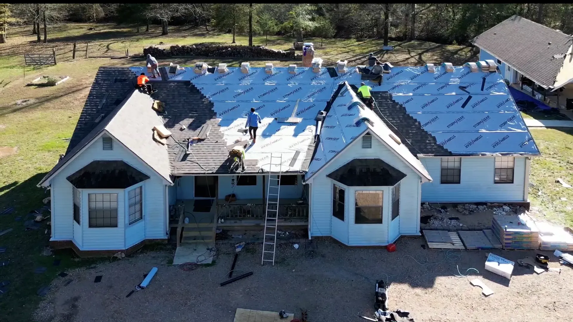 Crew installing new shingles over blue underlayment on a large white home on a wooded lot